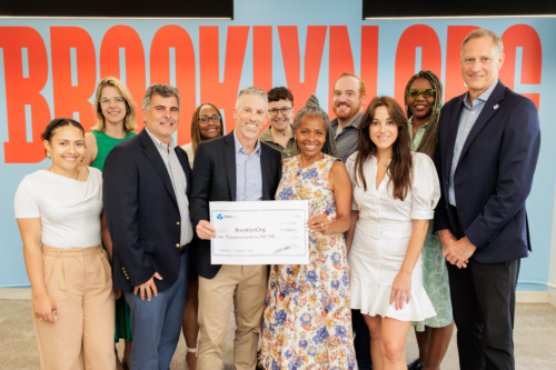 A group of people pose indoors in front of a “Brooklyn Org” sign, with two people holding a large check made out to BrooklynOrg for $10,000.