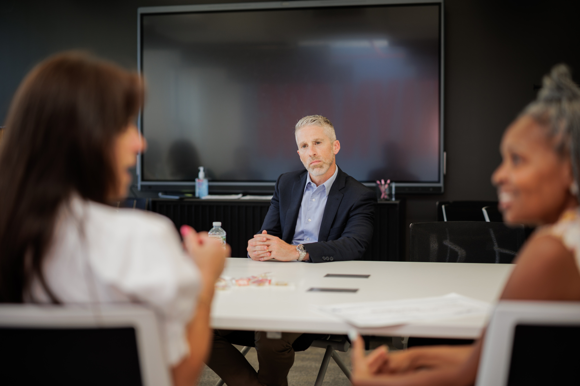 A man in a suit sits at a conference table, listening to two women talking, with a large monitor in the background.