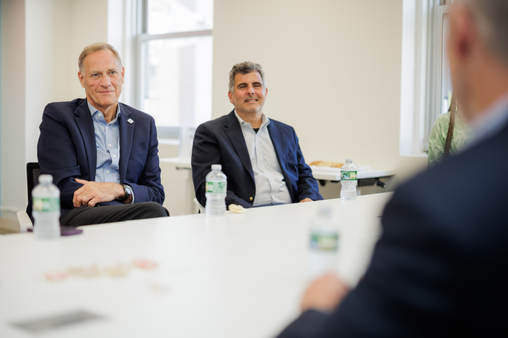 Two men in business attire sit at a conference table with bottled water, participating in a meeting.
