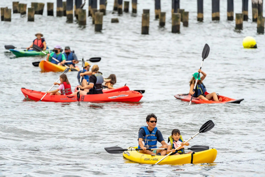 People kayaking in groups on a body of water, wearing life jackets. Some are in pairs, including adults and children. Wooden posts and a floating buoy are visible in the background.