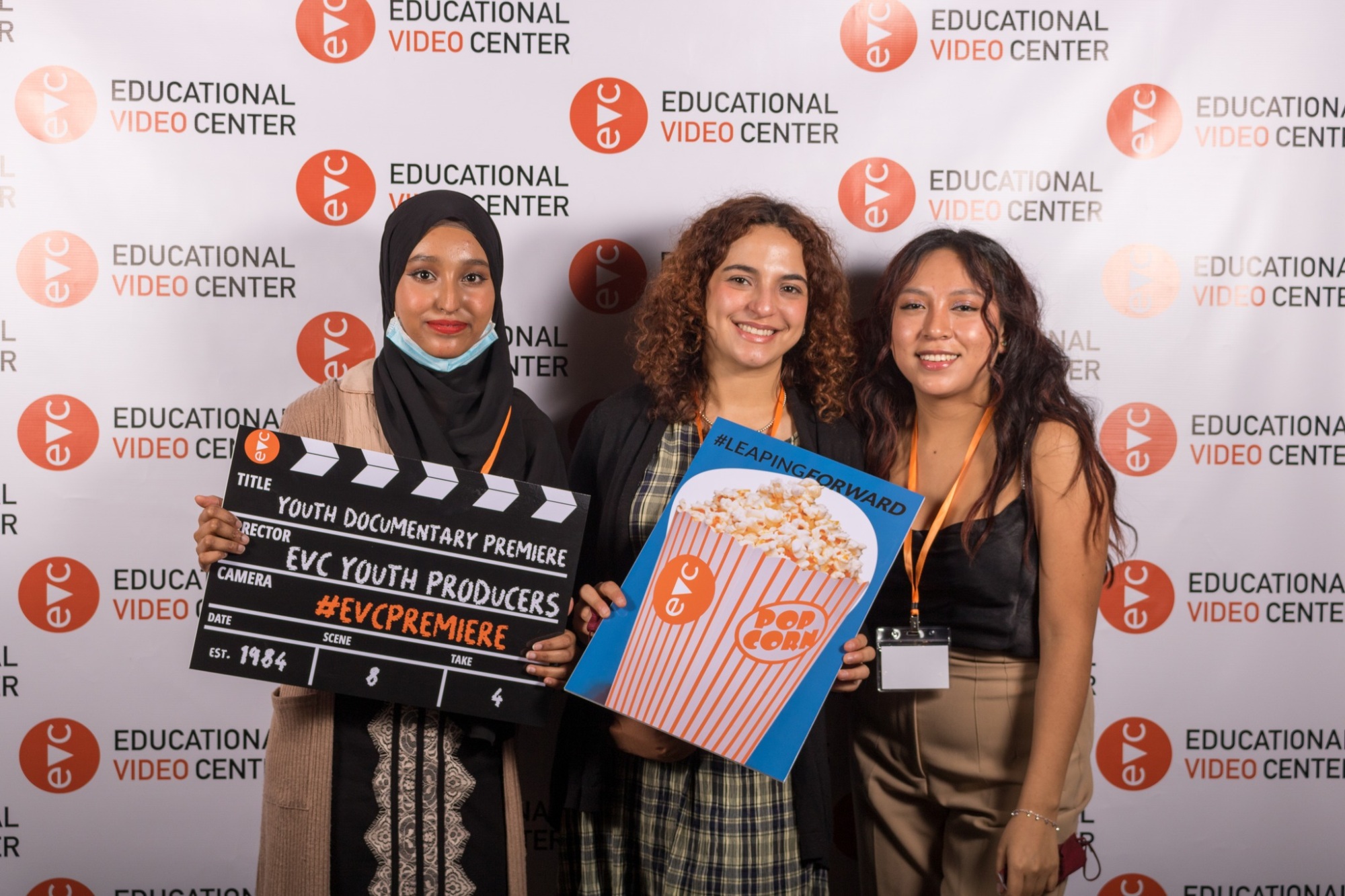 Three young women stand in front of an Educational Video Center backdrop, holding a film clapperboard and a popcorn sign, smiling for the camera.