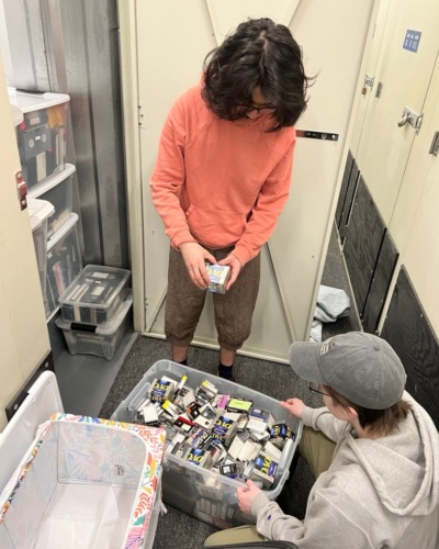 Two people sorting and examining trading cards from a plastic storage bin in a room filled with shelves and cabinets.