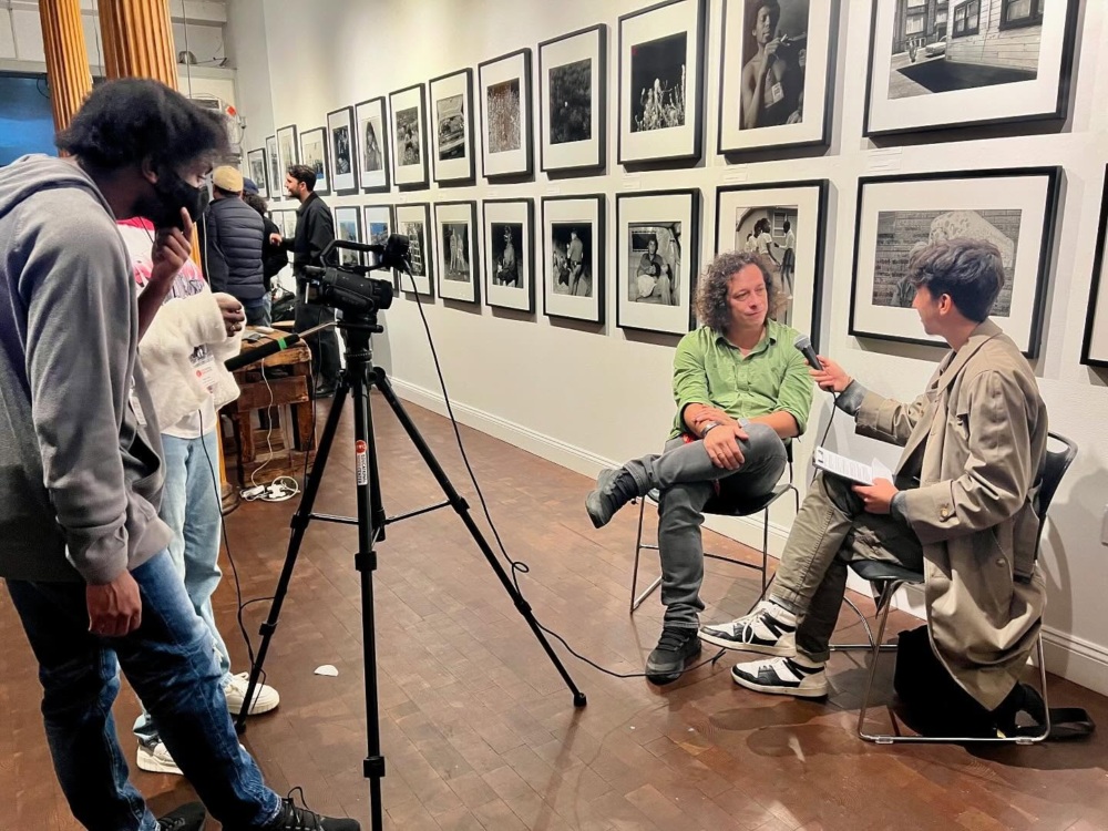 Two people sit in front of a camera for an interview in an art gallery, while two other people manage filming equipment. Black-and-white photographs hang on the walls behind them.