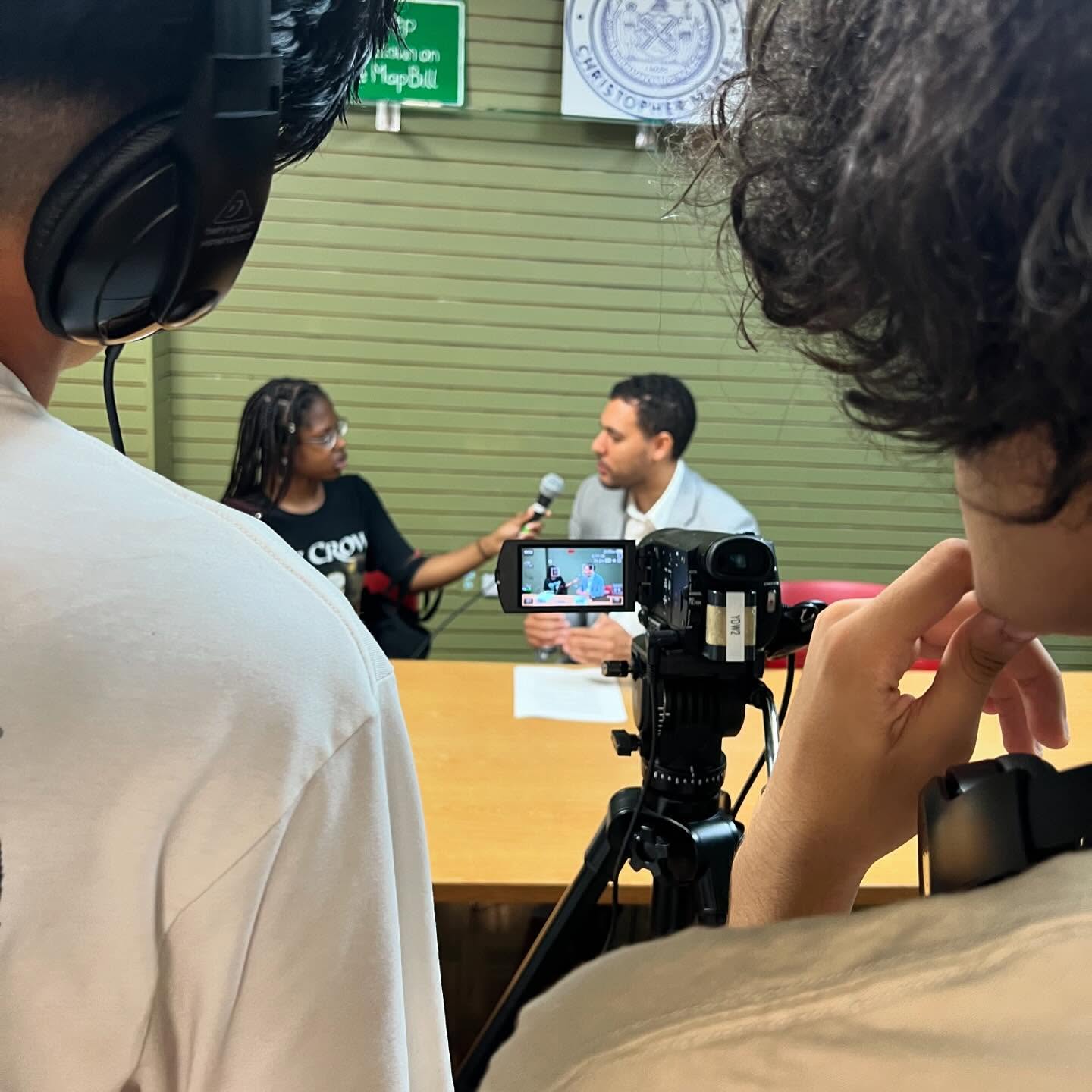 Two people are recording an interview at a table while a camera films them. Two crew members with headphones are visible in the foreground.