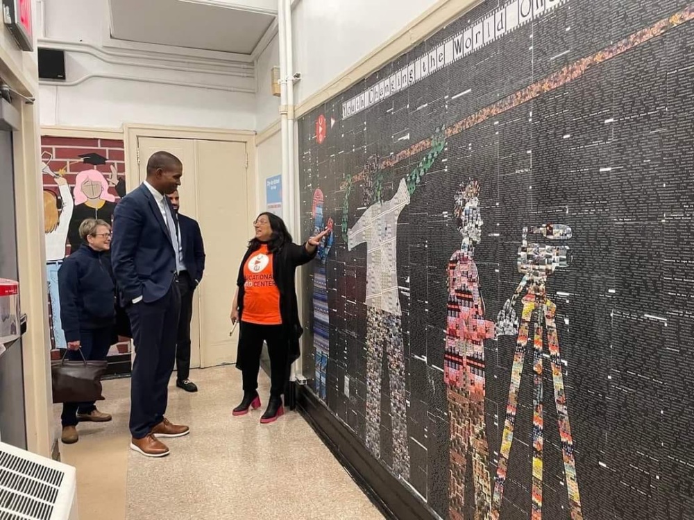 Four adults stand in a hallway, observing a large mosaic mural on the wall while one woman gestures toward the artwork.