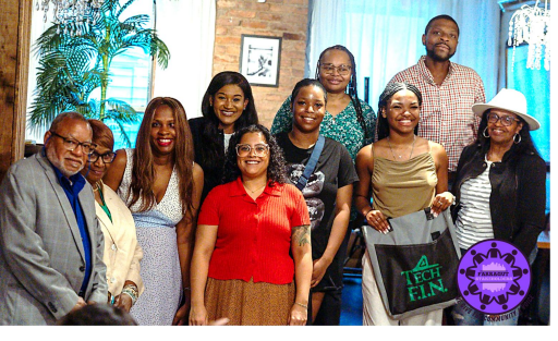 A group of ten adults pose and smile for a photo indoors; some are standing, others seated. A purple logo with people holding hands is in the bottom right corner.