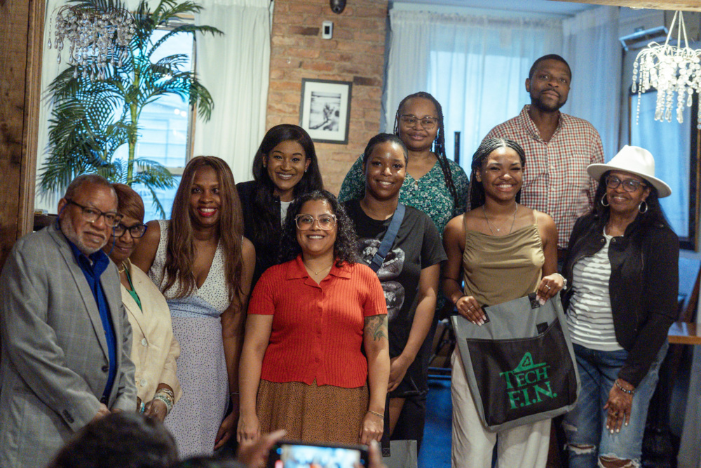 A group of ten people, smiling and posing indoors in front of a window, with one person holding a "Tech F.I.N." bag.
