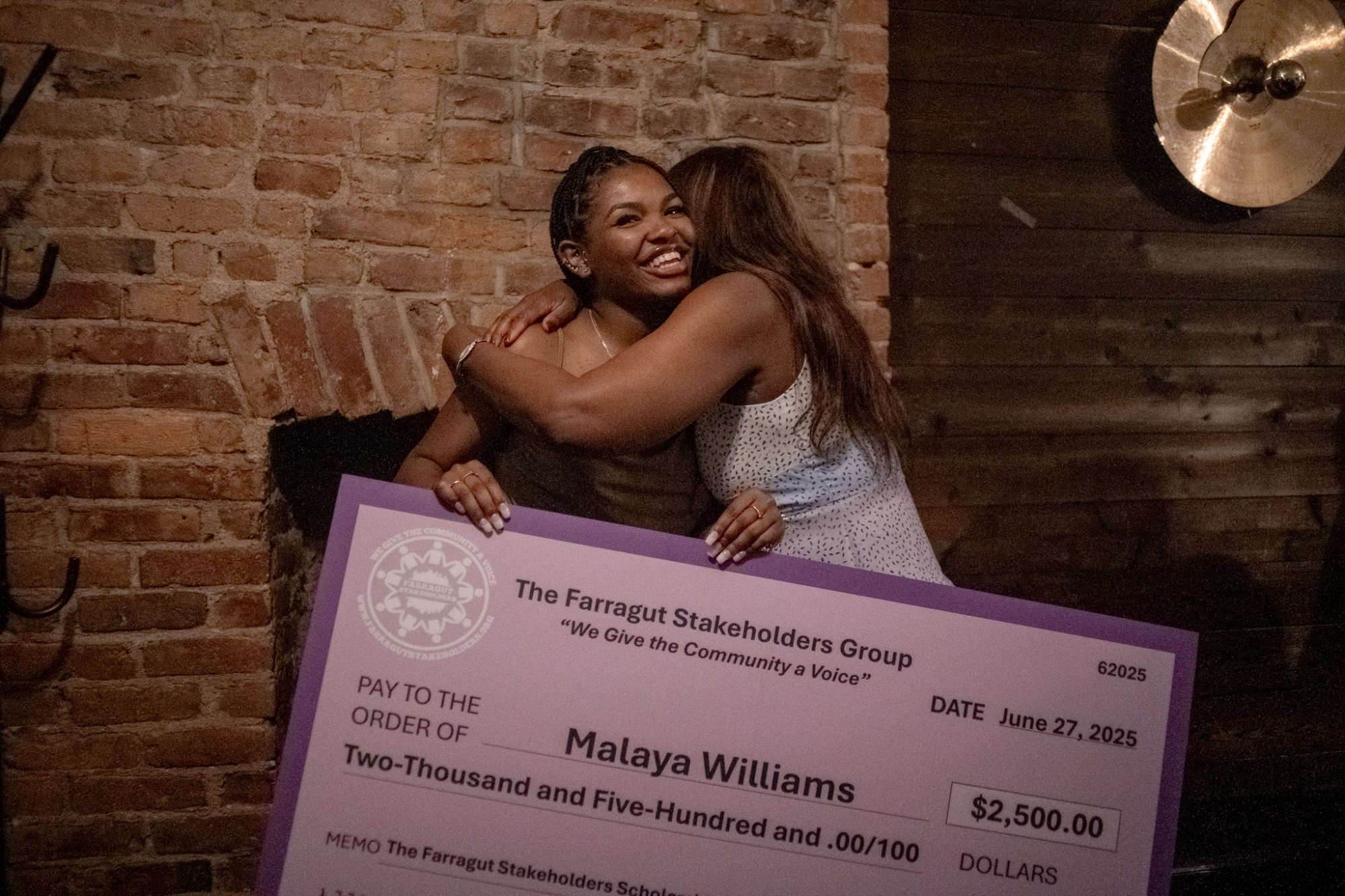 Two women embrace while holding a large ceremonial check for $2,500 made out to Malaya Williams from The Farragut Stakeholders Group, dated June 27, 2025.