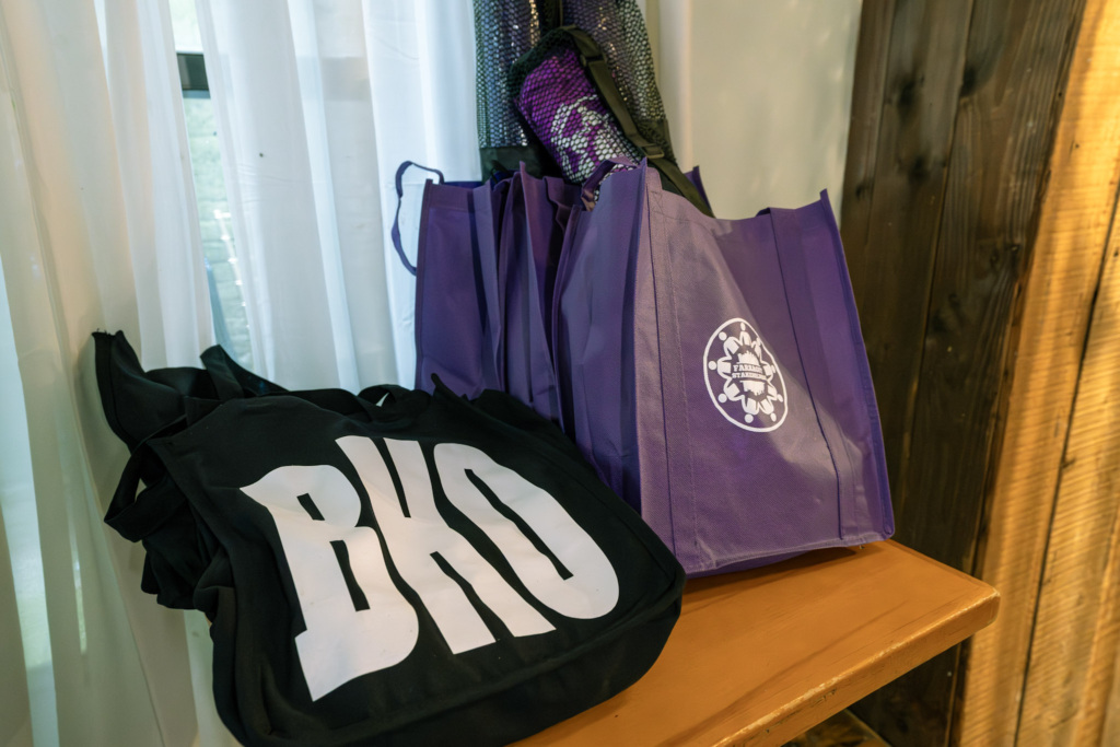 A black tote bag with large white letters "BKO" is next to several purple tote bags with a white circular logo, all placed on a wooden surface by a window.