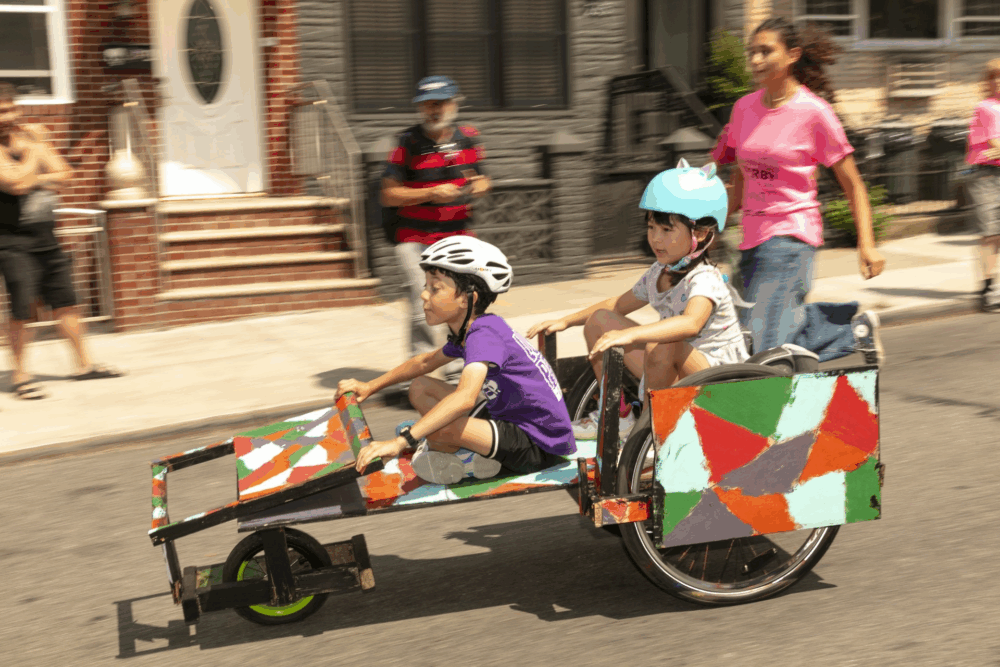 Two children wearing helmets ride a colorful, homemade go-kart on a street, while an adult and another person watch and walk alongside.