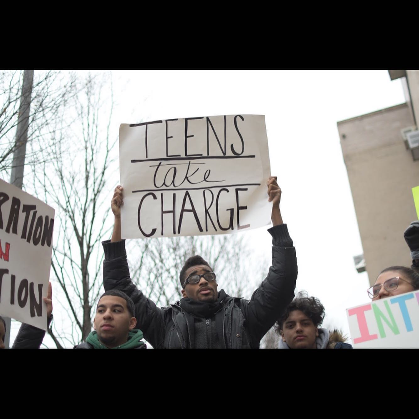 A group of young people stand outdoors; one person in the center holds a sign reading "TEENS take CHARGE.