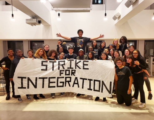 A group of diverse students pose indoors holding a large banner that reads "STRIKE FOR INTEGRATION.
