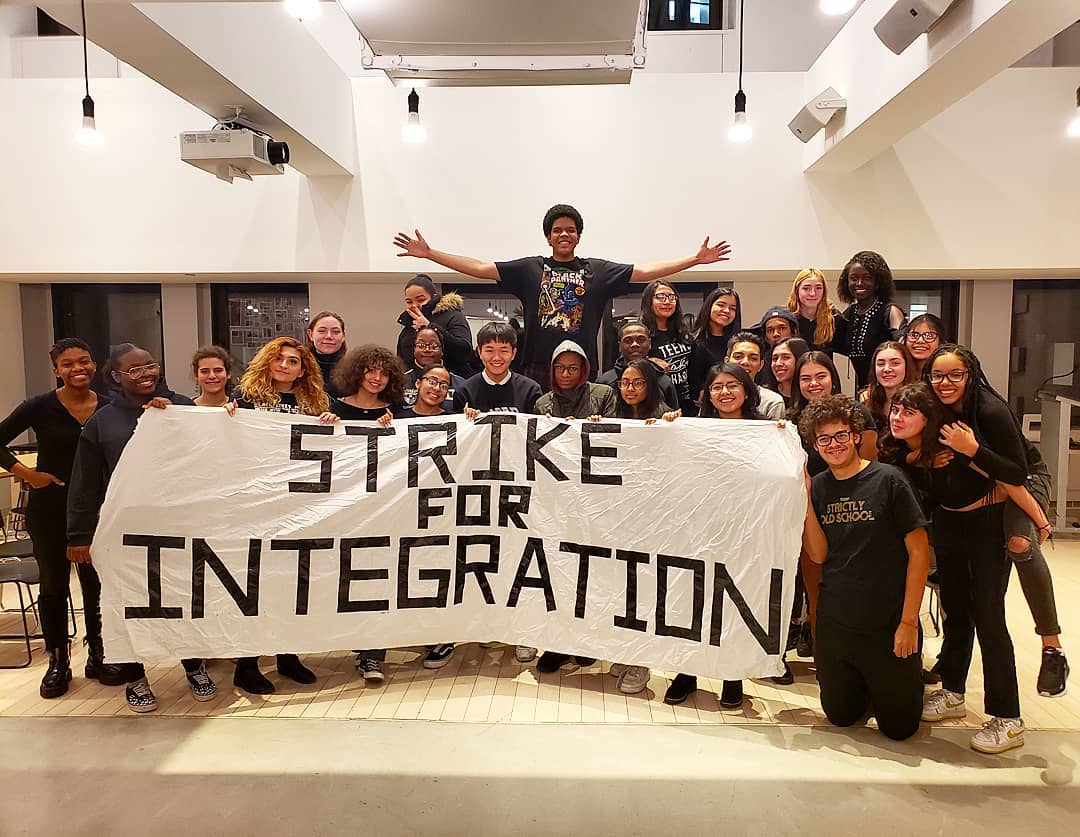 A group of diverse students pose indoors holding a large banner that reads "STRIKE FOR INTEGRATION.