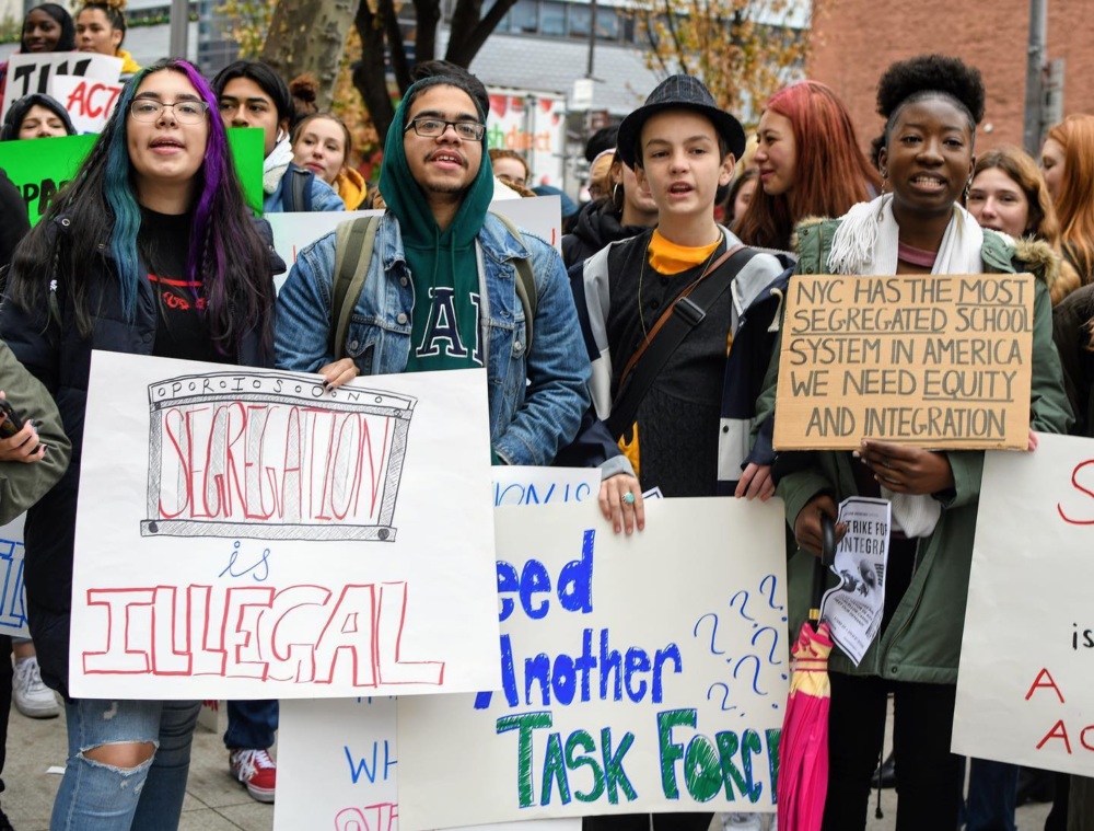 A diverse group of young people hold signs protesting school segregation and calling for equity and integration at a rally.