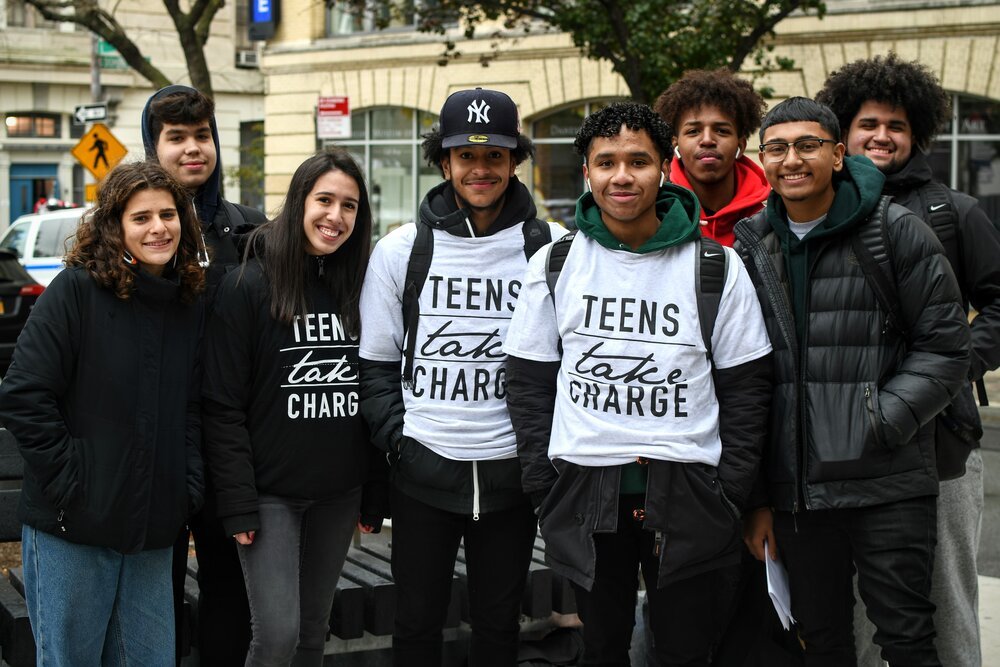 A group of eight teenagers, some wearing "Teens Take Charge" shirts, stand together outdoors and smile at the camera.