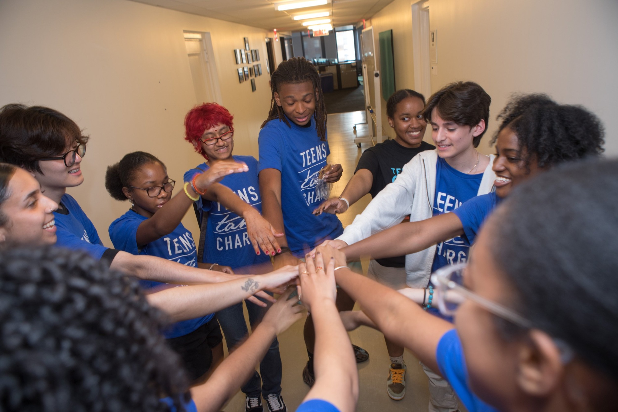 A group of teenagers and an adult stand in a circle in a hallway, placing their hands together in the center as a gesture of teamwork.