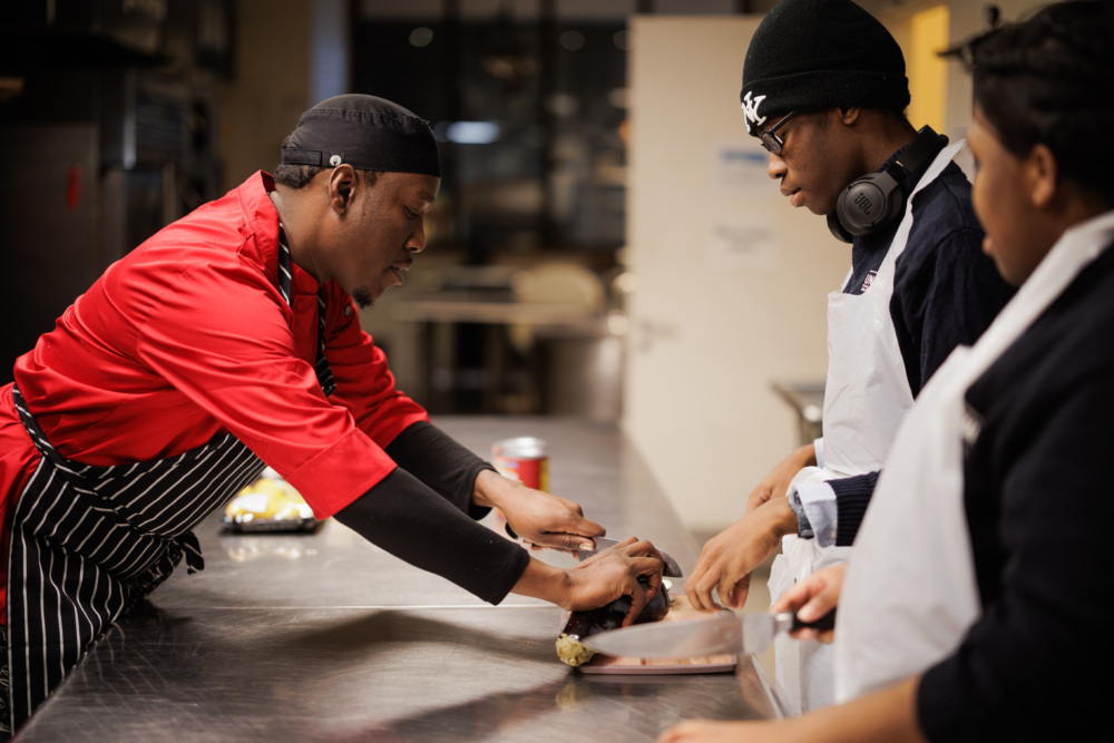 A chef in a red uniform demonstrates food preparation to two students at a stainless steel kitchen counter.