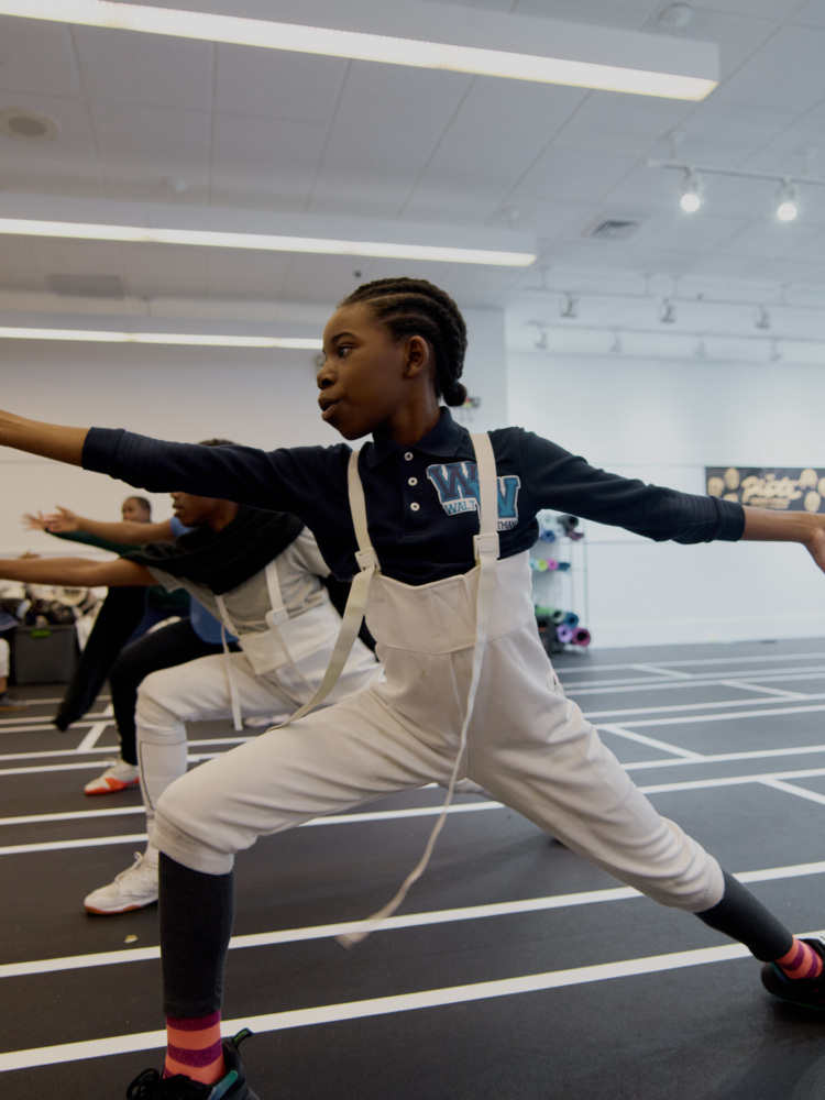 A child in sportswear practices a fencing stance in a brightly lit indoor gym, with others in similar attire behind them.