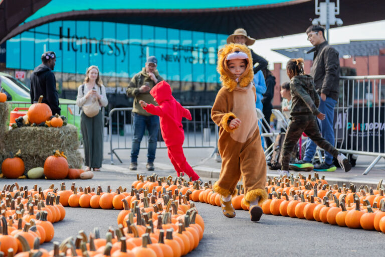 A child in a lion costume walks through a path lined with pumpkins at an outdoor event, with other children and adults in the background.