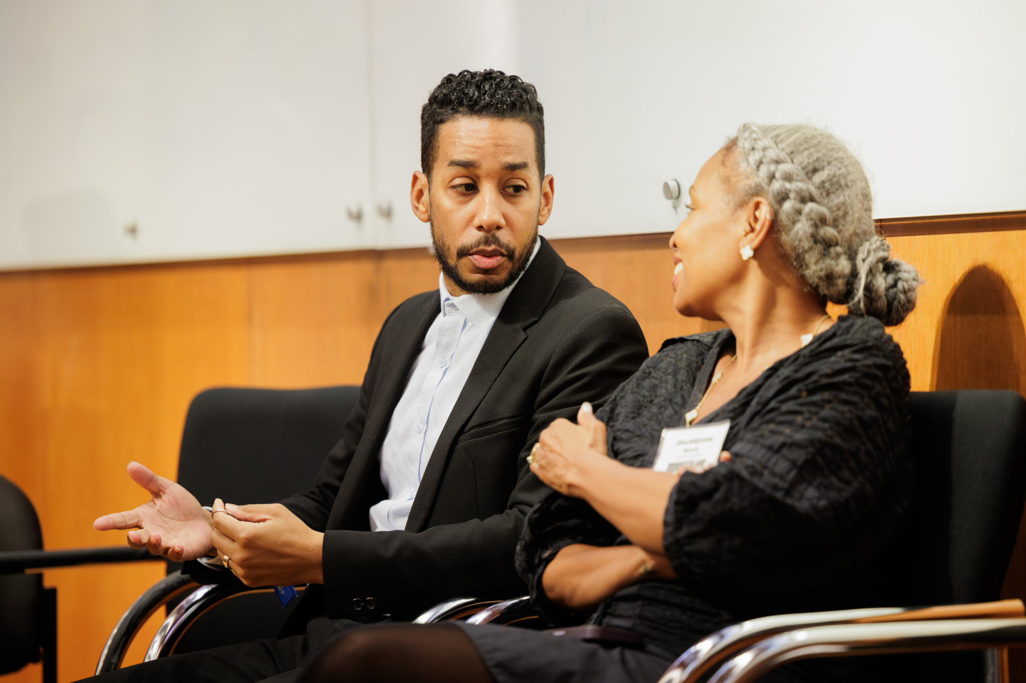 Two people sit on chairs in conversation, one gesturing with his hands while the other listens with arms crossed. Both wear business attire in an indoor setting.