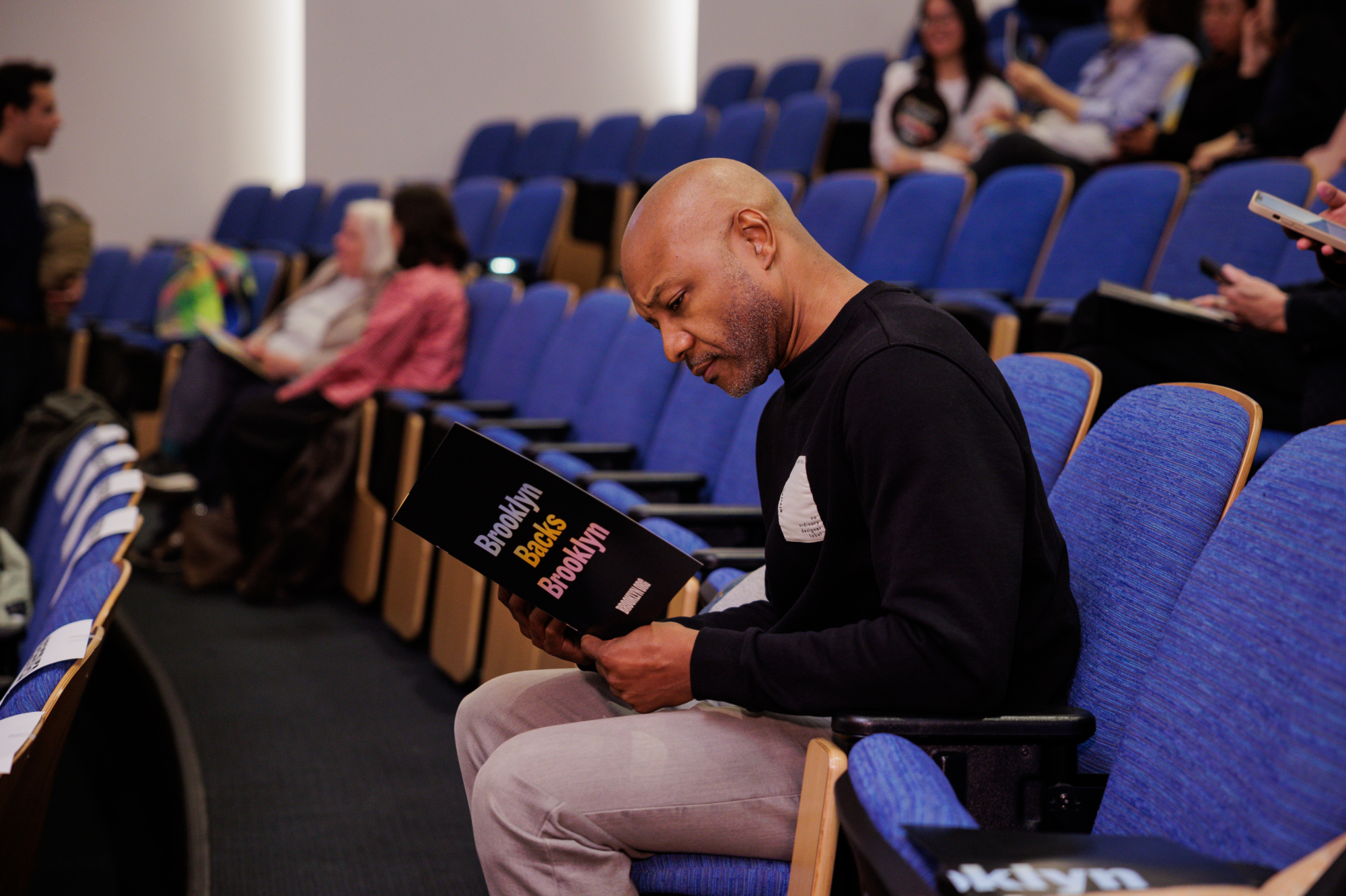 A man sits alone in a row of blue auditorium seats, reading a black program labeled "Brooklyn Books Brooklyn." Other people are seated in the background.