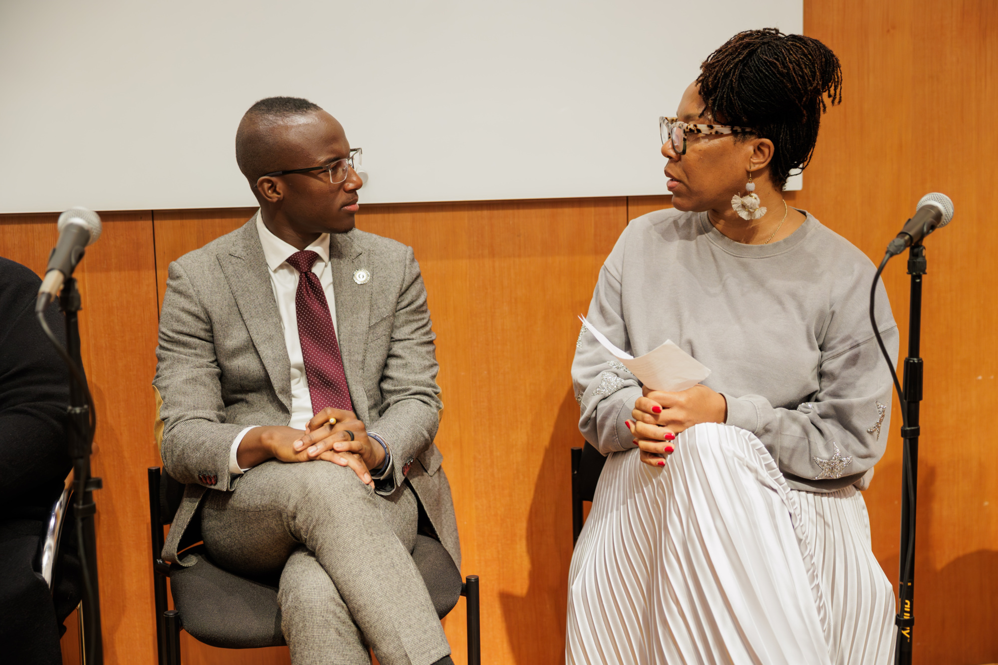 A man in a gray suit and a woman in a gray top and striped skirt sit on chairs, facing each other and talking, with microphones nearby.
