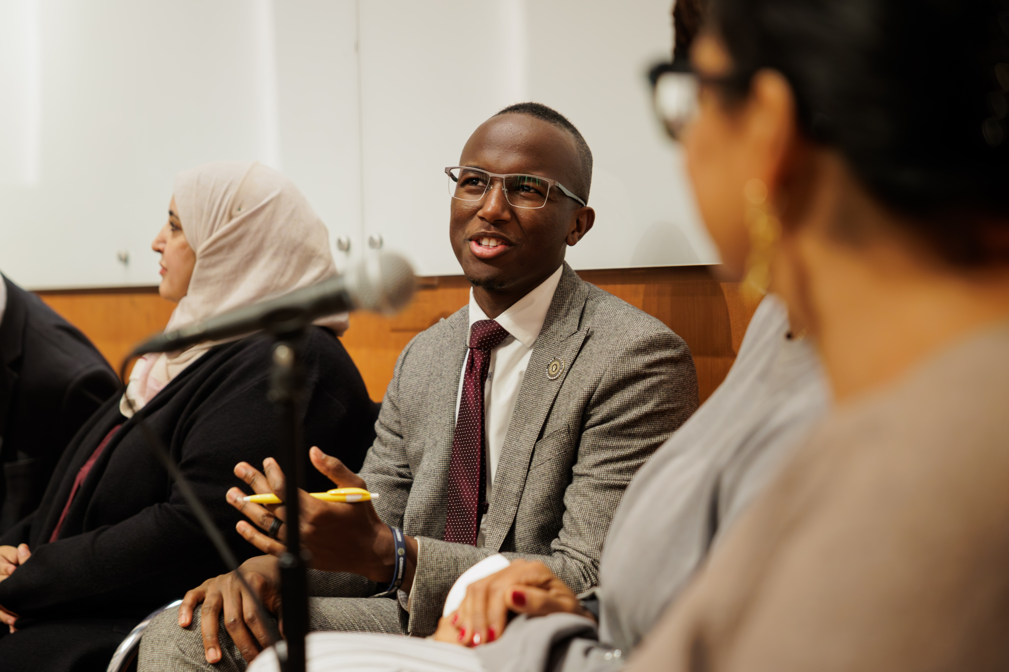 A man in a gray suit speaks and gestures during a panel discussion, with two women, one in a hijab, seated beside him and a microphone in the foreground.