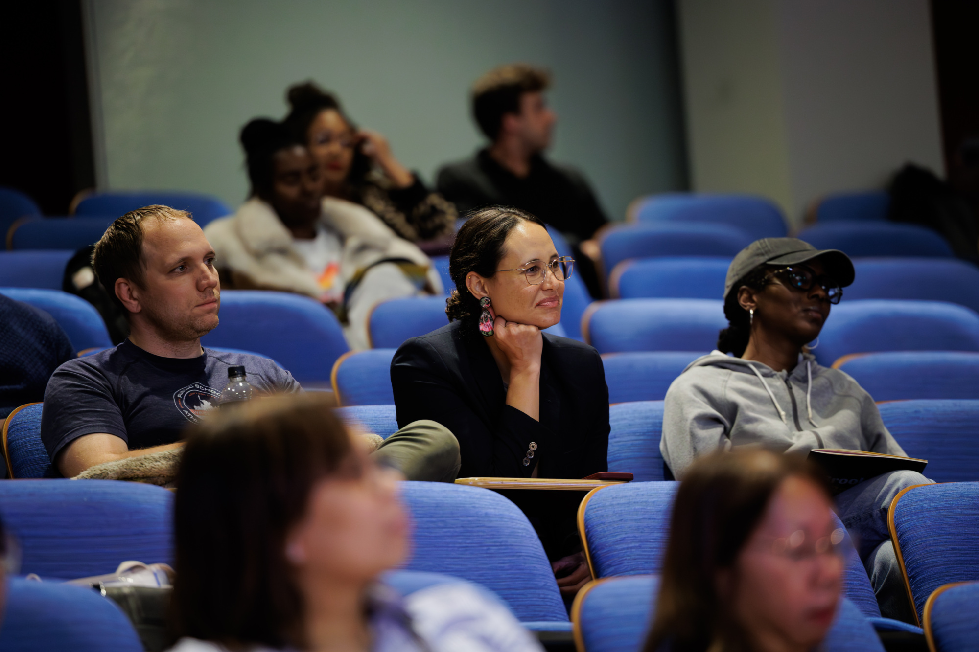 People sit in blue auditorium seats, attentively watching or listening to something off-camera. Some take notes while others observe quietly.