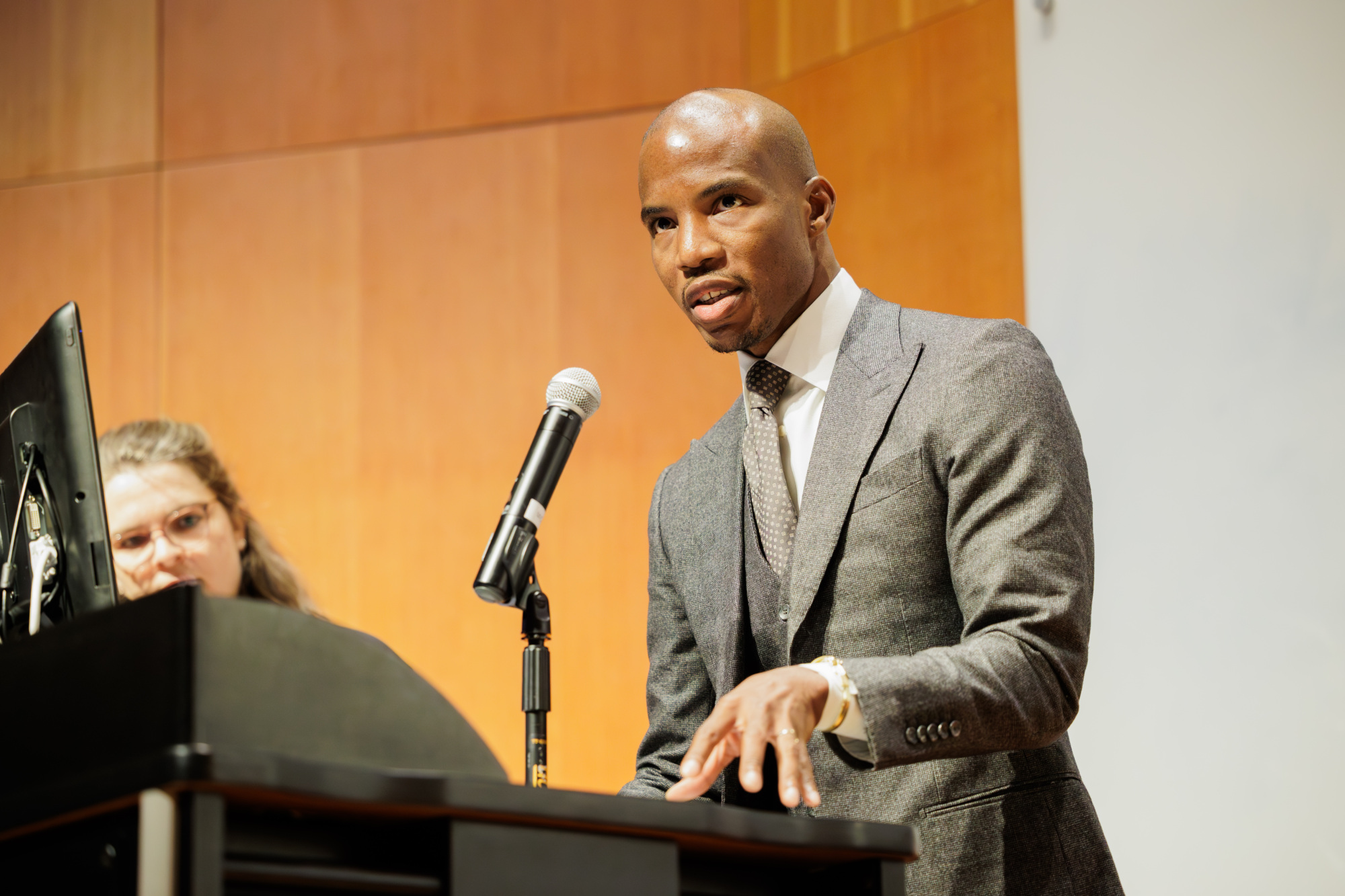 A man in a gray suit speaks at a podium with a microphone, while a woman stands beside him in a wood-paneled room.
