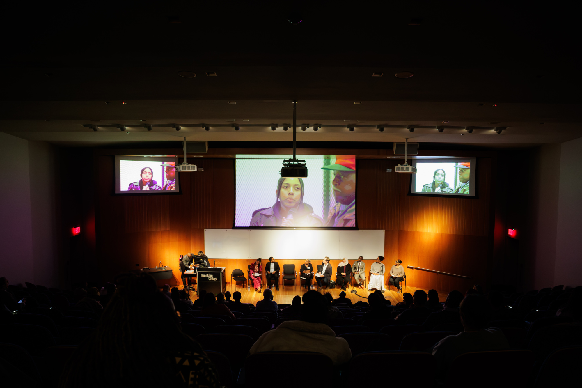 A panel of eight people sits onstage in an auditorium, speaking to an audience; the speaker's face is projected on screens behind them.