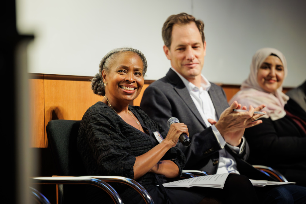 Three people sit on a panel; the woman in front holds a microphone and smiles, while the man and another woman sit beside her, with the man clapping.