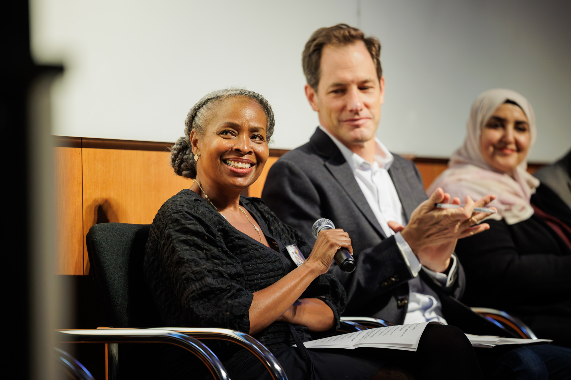 Three people sit on a panel; the woman in front holds a microphone and smiles, while the man and another woman sit beside her, with the man clapping.