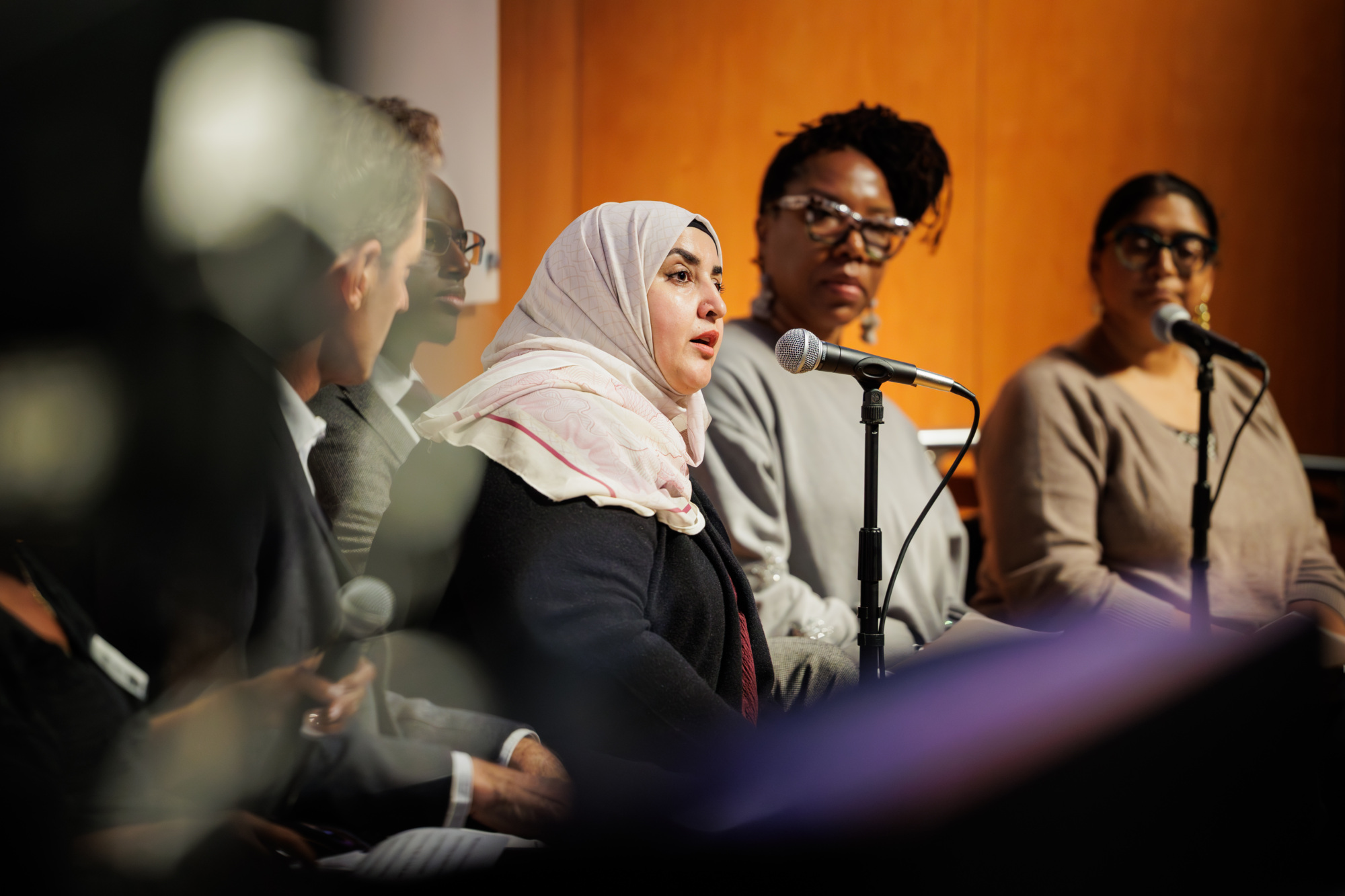A woman in a hijab speaks into a microphone during a panel discussion, with three other people seated beside her, all holding microphones.