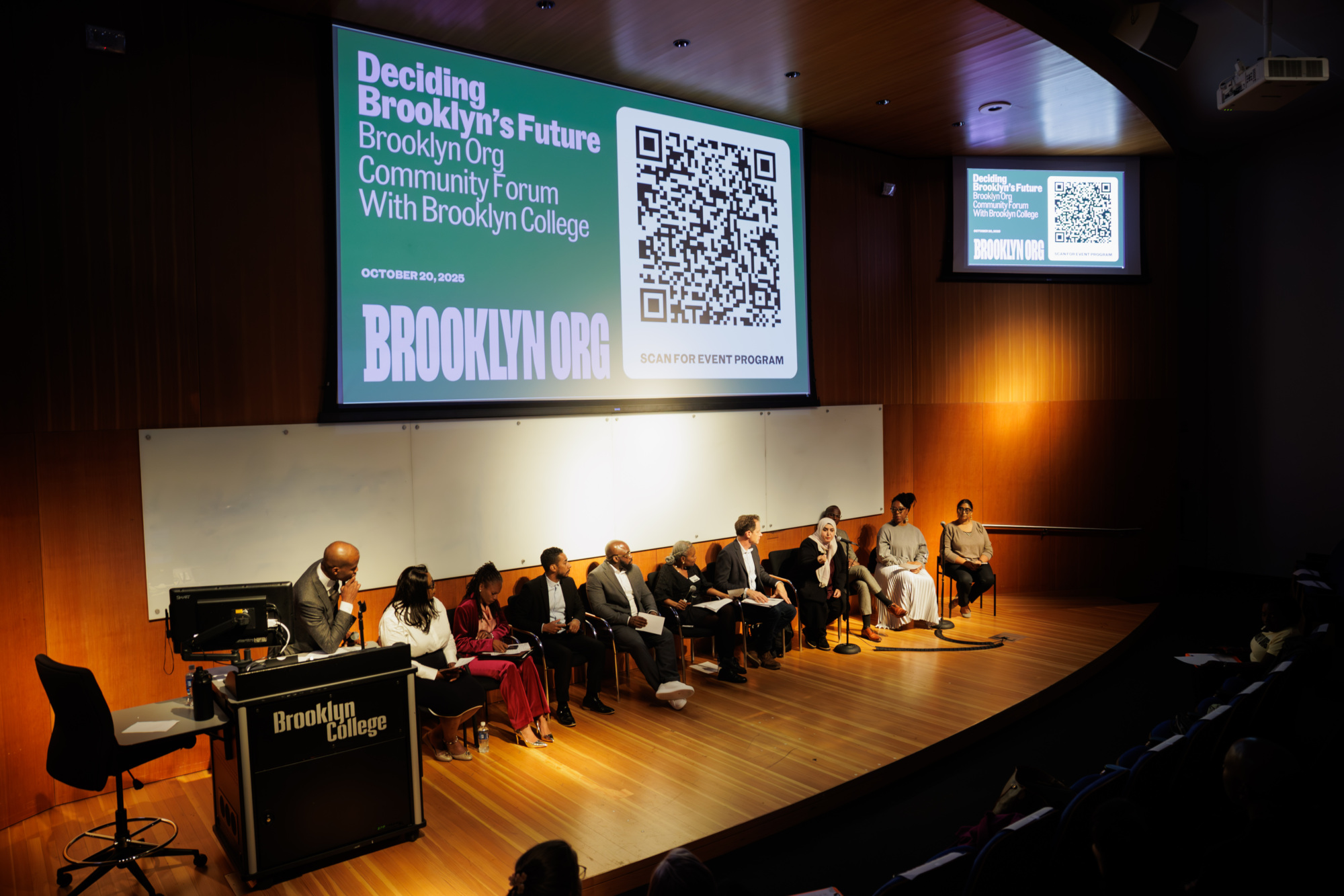A panel of speakers sits on stage during a community forum at Brooklyn College, with event details and a QR code displayed on large screens behind them.