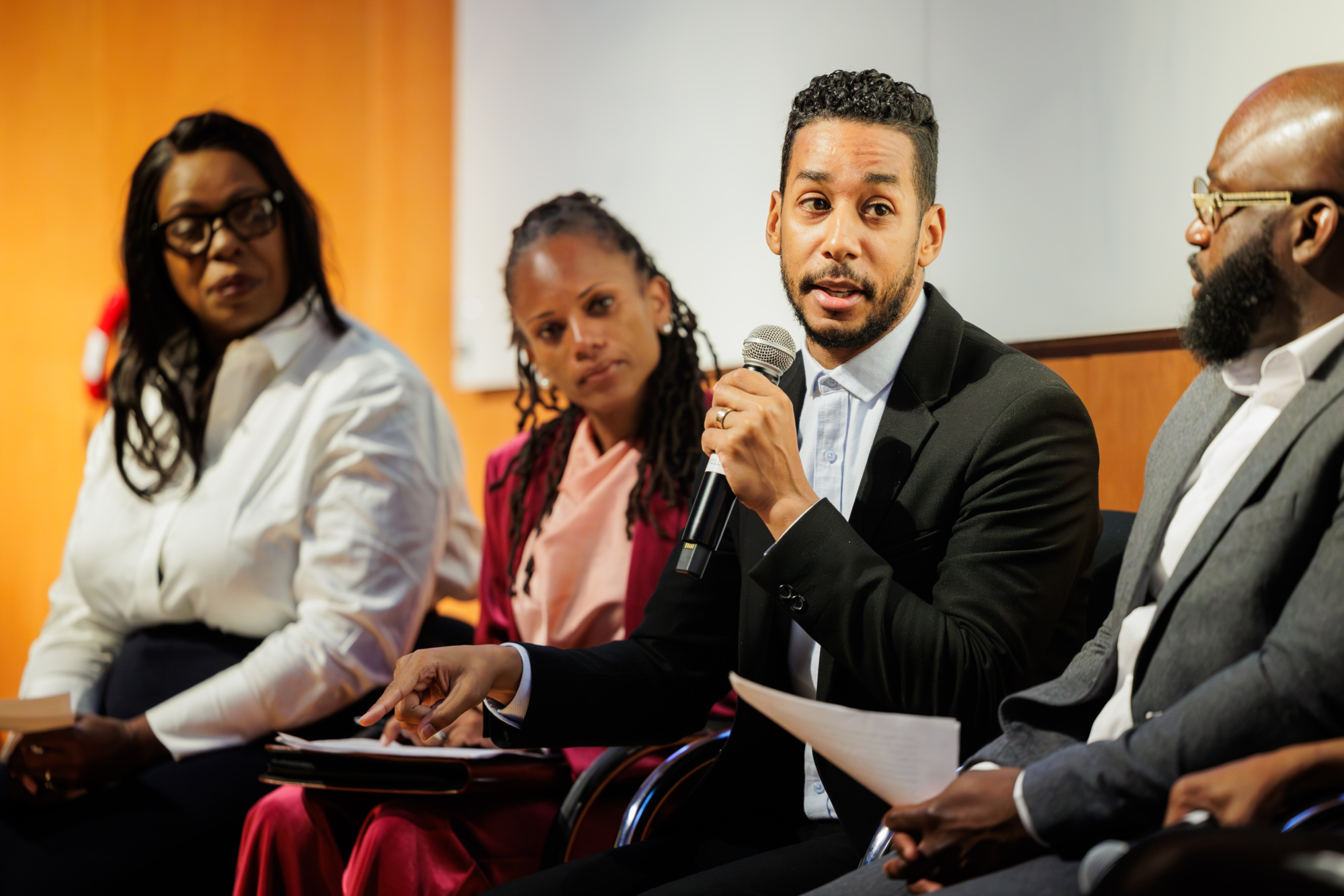 A man in a suit speaks into a microphone while seated on a panel with three other people, two women and one man, during an indoor event.