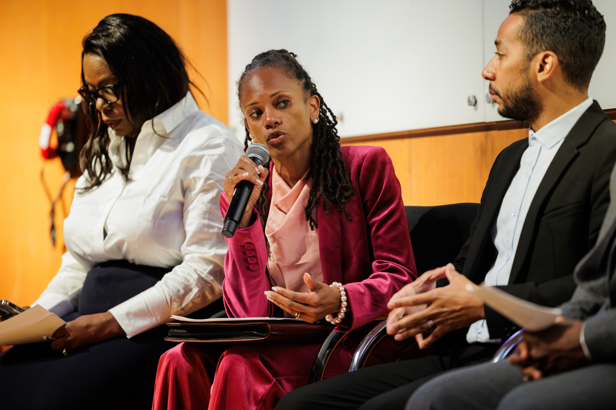Three people sit on a panel; the woman in the center, wearing a pink suit, speaks into a microphone while the others listen.