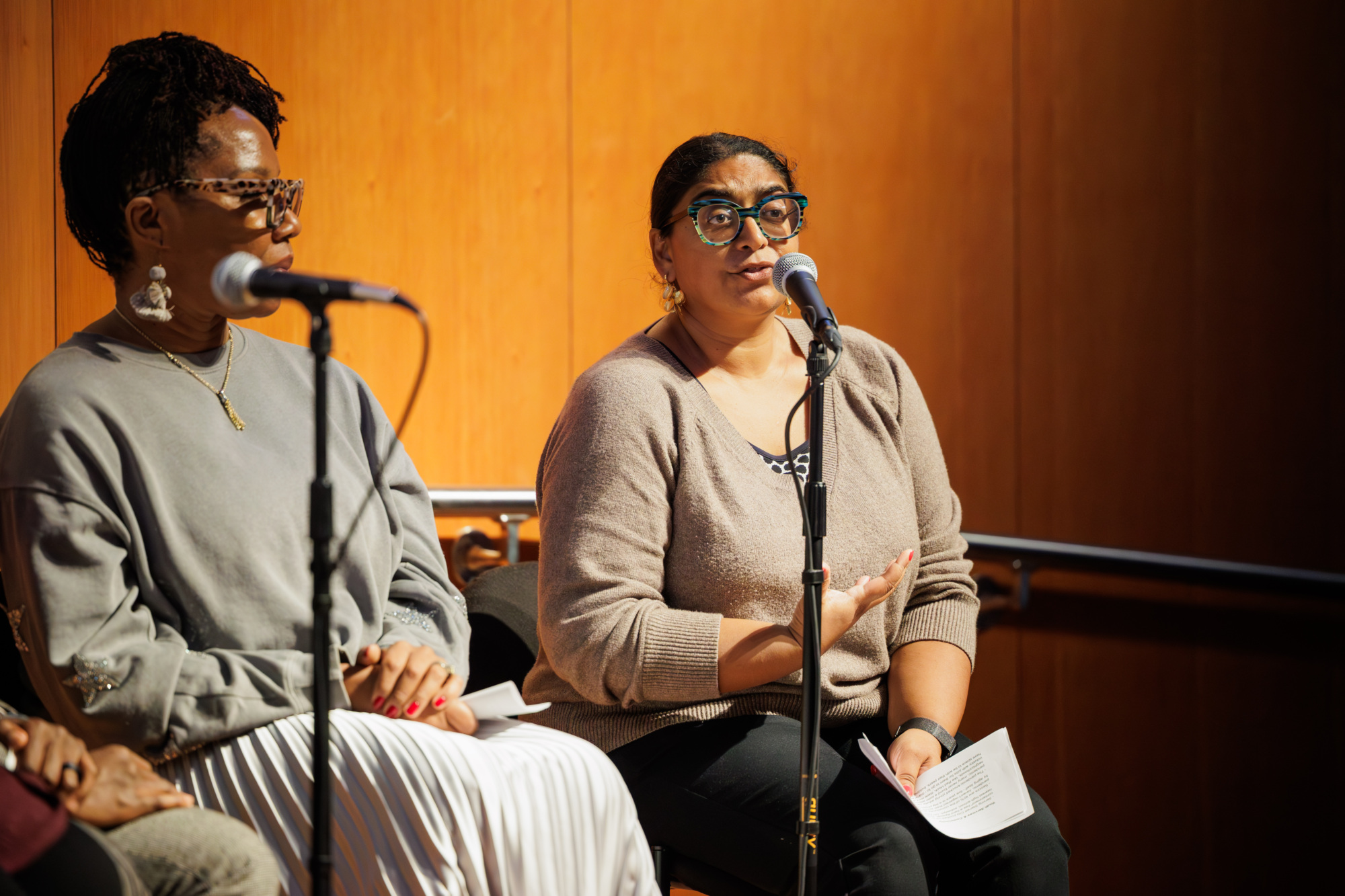 Two women sit on a stage, each speaking into a microphone; one gestures with her hand while holding papers, the other listens attentively.