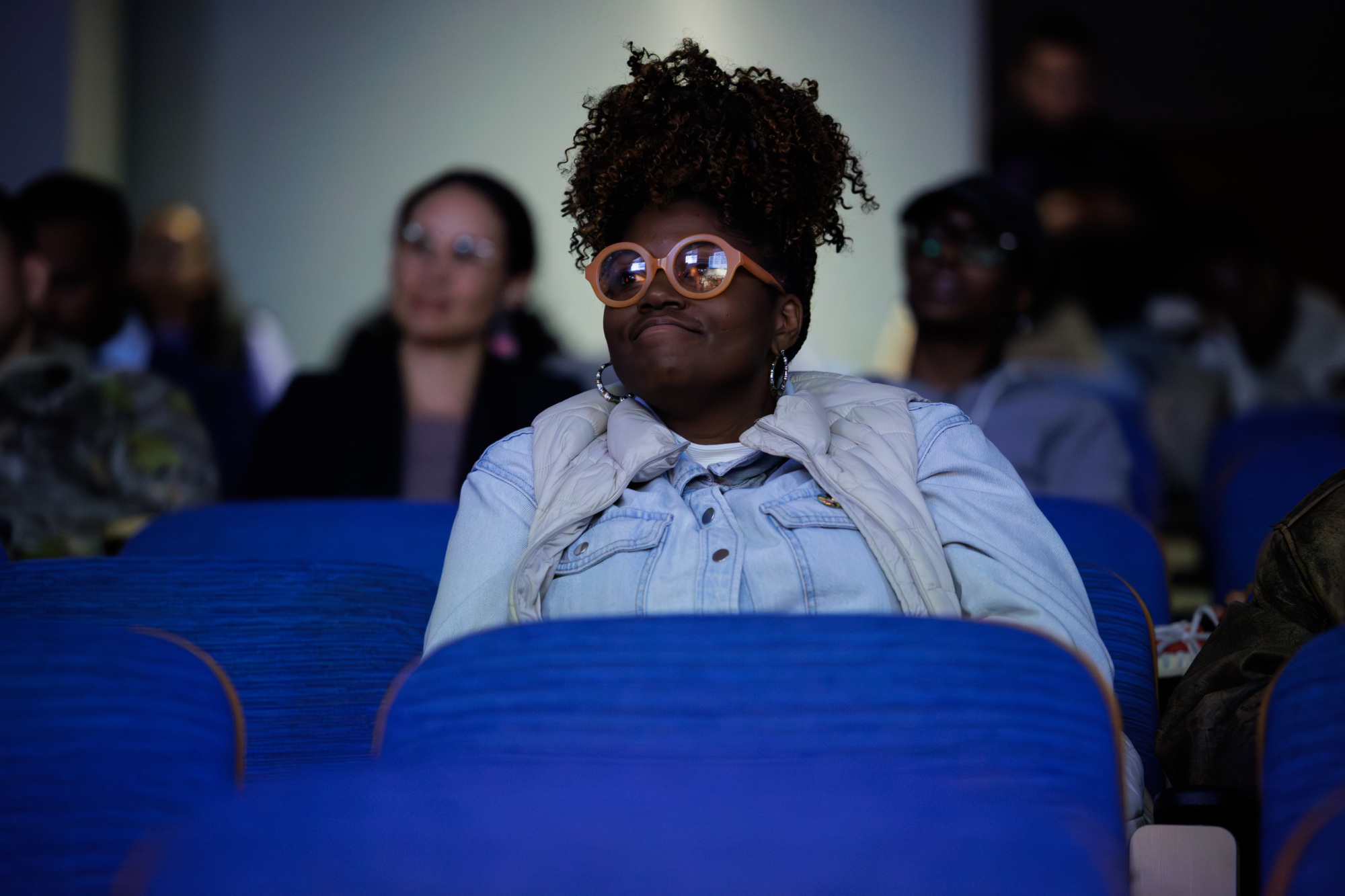 A person wearing glasses and a light-colored jacket sits alone in a row of blue auditorium seats, with other people blurred in the background.