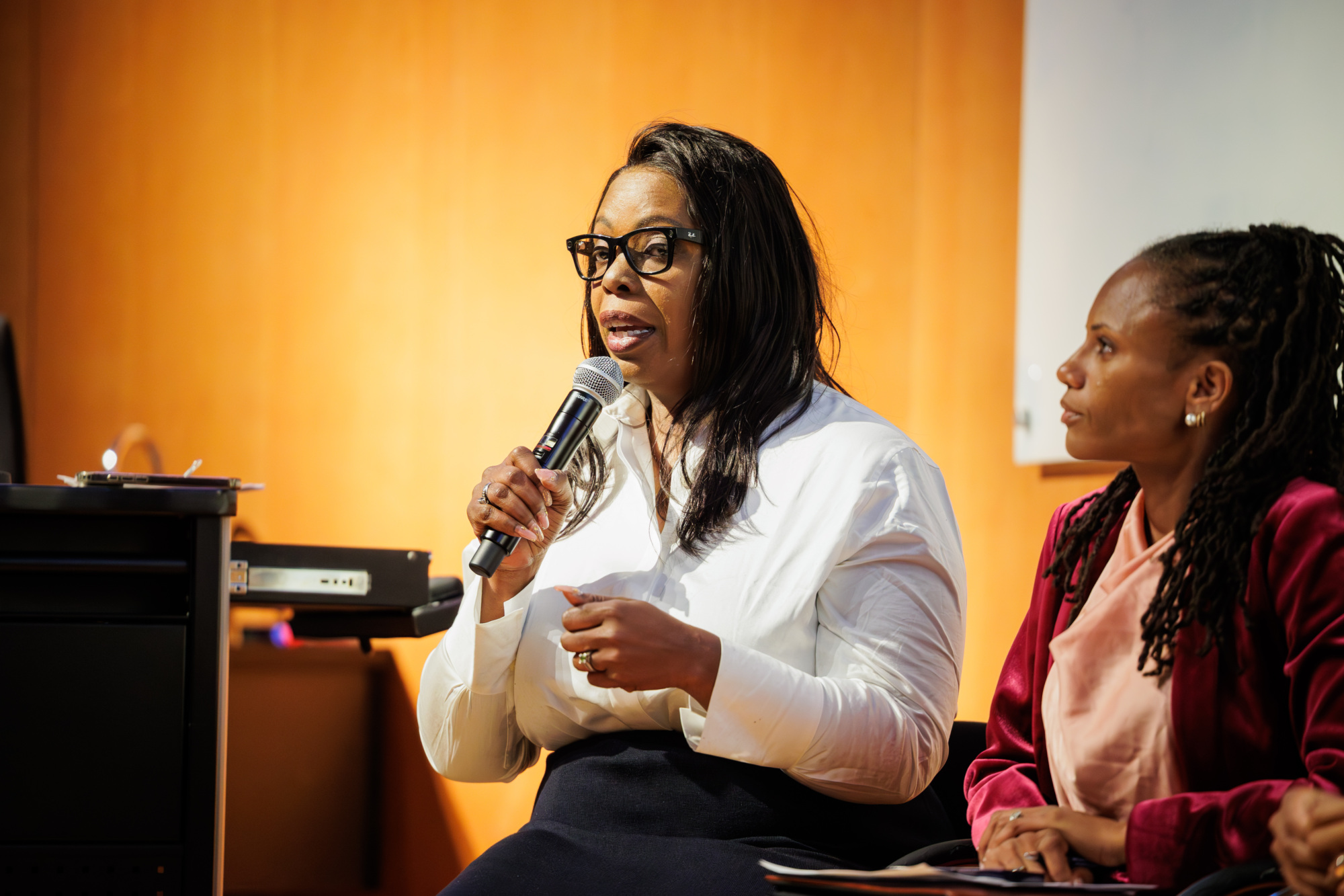 Two women sit indoors; one is speaking into a microphone while the other listens attentively, holding papers in her lap.