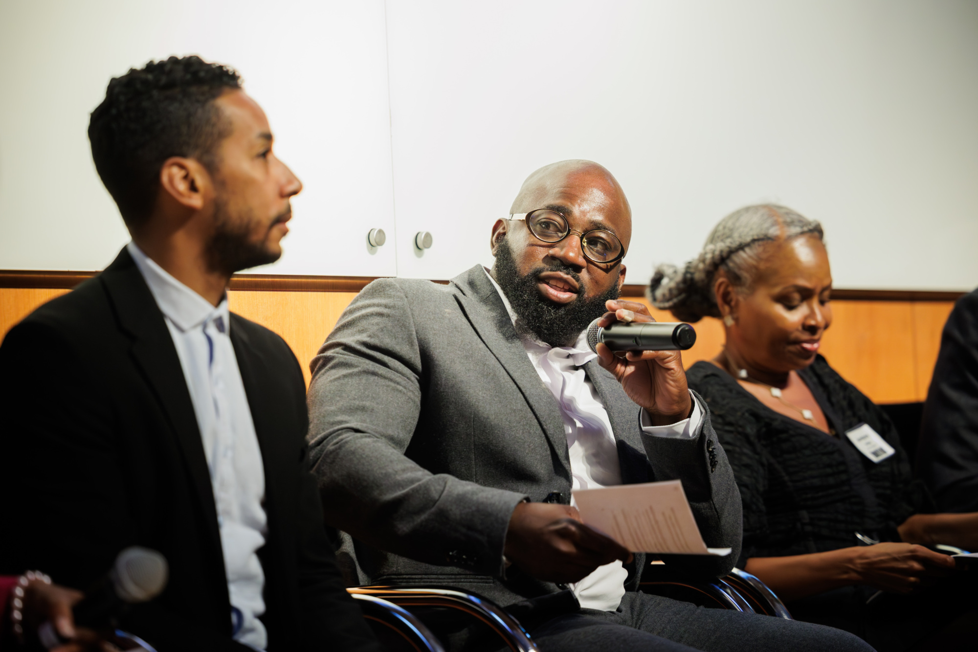 Three people sit on a panel, with the man in the center holding a microphone and speaking, while the others listen attentively.