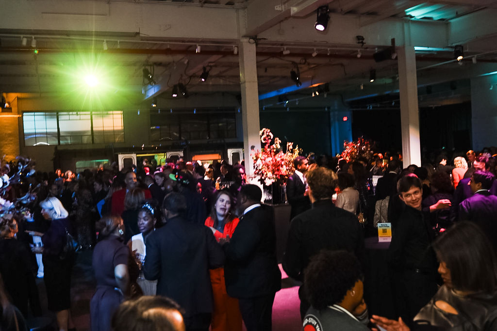 A large crowd of people socializing at an indoor event space with colorful lighting and floral centerpieces.