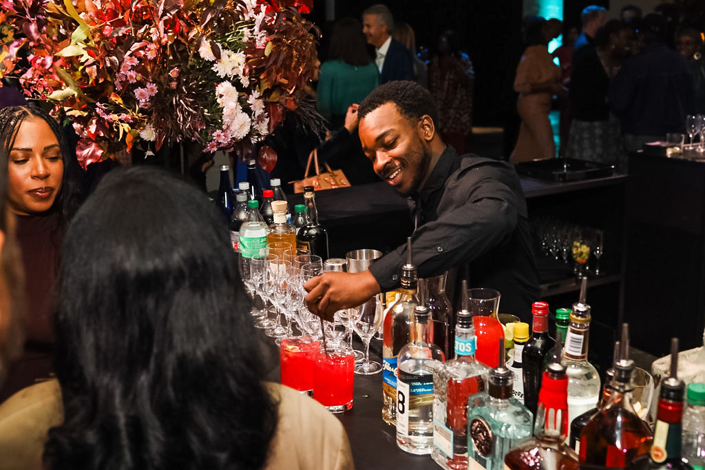 A bartender is preparing drinks behind a bar with assorted bottles, while people gather and socialize in a dimly lit setting.