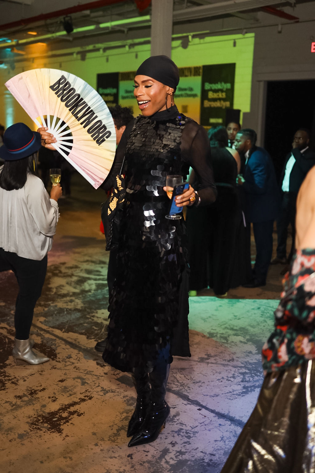 A person in a black dress holds a large fan reading "BROOKLYNWORDS" at an indoor social event with other attendees in the background.