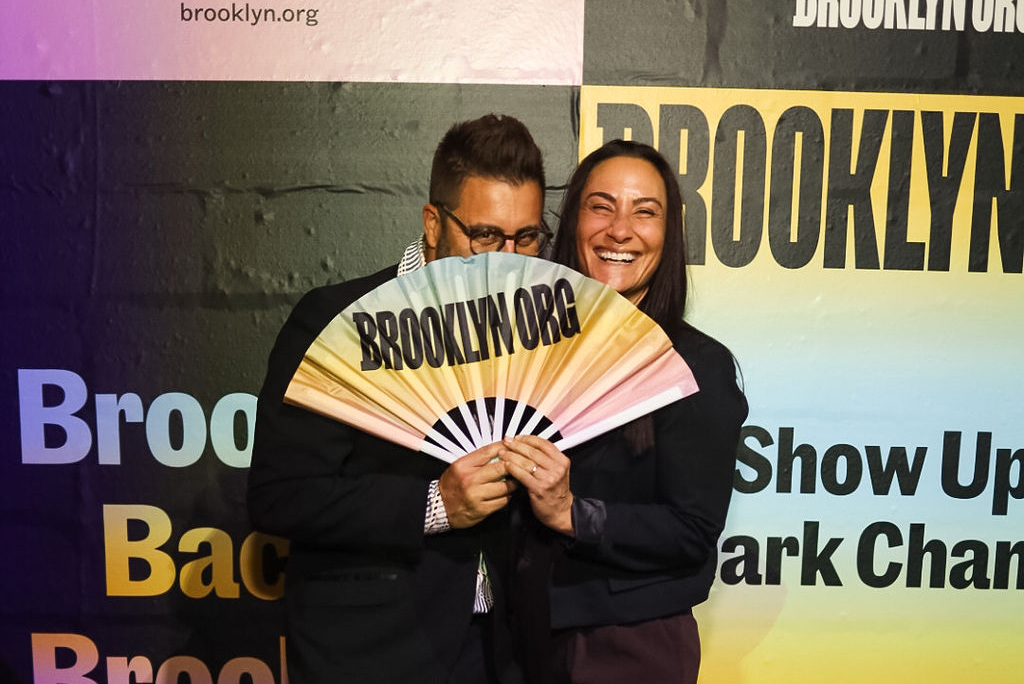 Two people stand smiling in front of a "Brooklyn.org" backdrop, partially covering their faces with a large fan that reads "BROOKLYN.ORG.