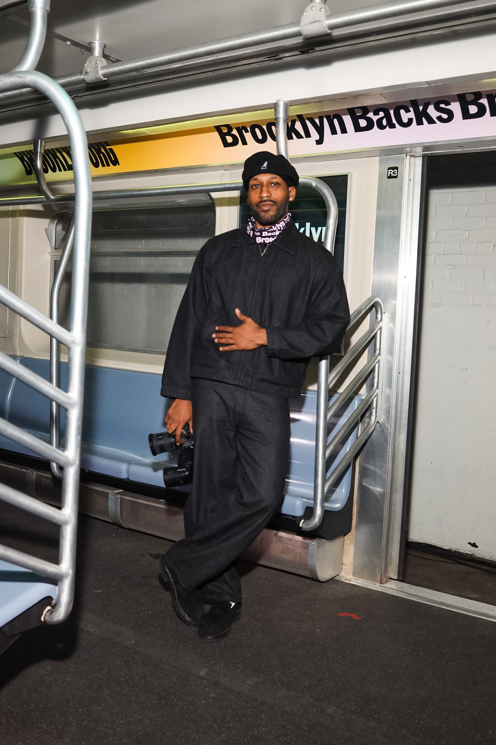 A man wearing dark clothing and a black beret stands in a subway car, holding a camera with one hand and resting the other on his stomach. The sign above him reads "Brooklyn Backs.