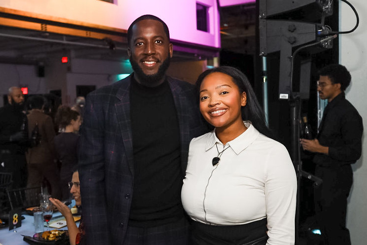 A man in a plaid suit and a woman in a white blouse and black skirt stand and smile at a formal event with seated guests in the background.