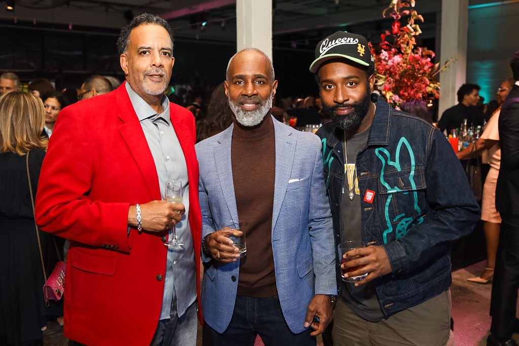 Three men pose together at an indoor social event, each holding a drink. The background shows other guests and floral arrangements.