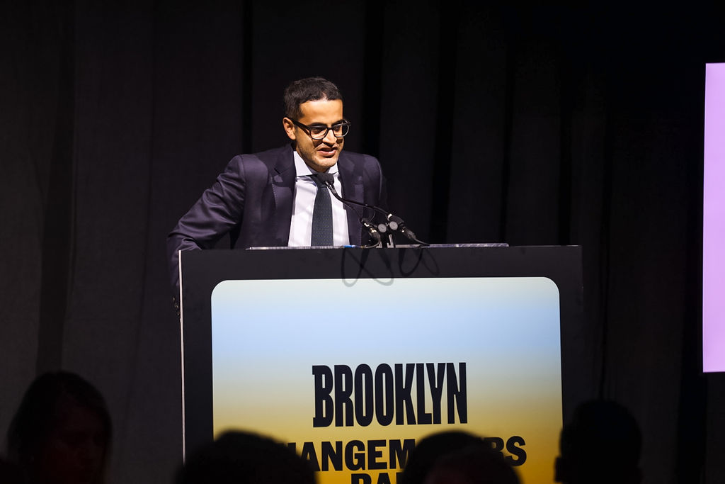 A man in a suit speaks at a podium with a sign that reads "Brooklyn" at an indoor event.