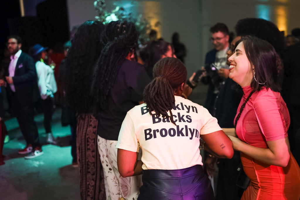 A group of people gather indoors at an event; one person wears a shirt reading "Brooklyn Backs Brooklyn" while others stand and talk nearby.