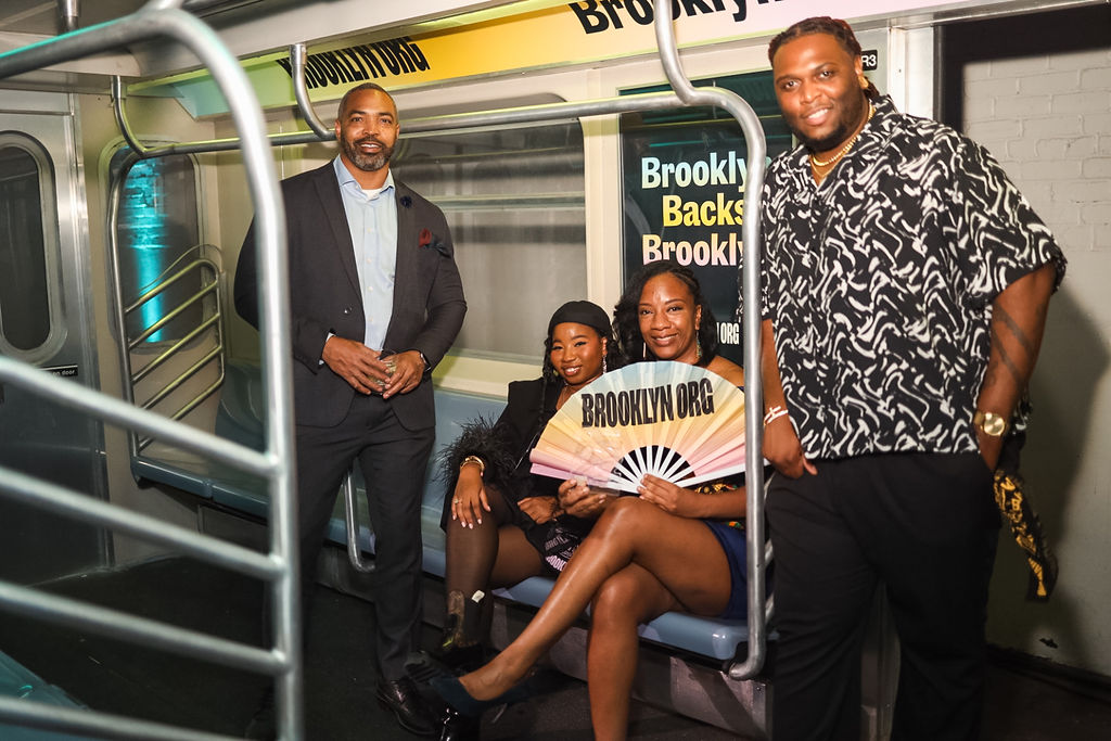 Four people pose and smile inside a subway car; two are seated, one holds a “BROOKLYN.ORG” fan, and two stand nearby. Signs promoting Brooklyn are visible in the background.